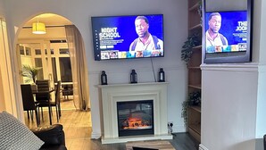 Een woonkamer met een open haard, een aan de muur bevestigde televisie, een bank en een zichtbare eethoek via een boogvormige doorgang.