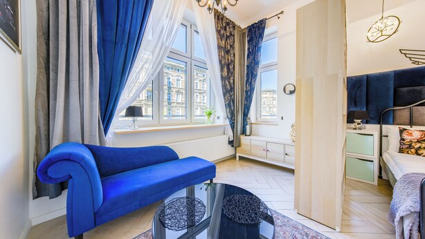 Ein modernes Schlafzimmer mit einem blauen Sofa, einem Bett mit einem blumigen Kopfkissen, einem runden Glastisch und Blick auf Gebäude durch das Fenster.