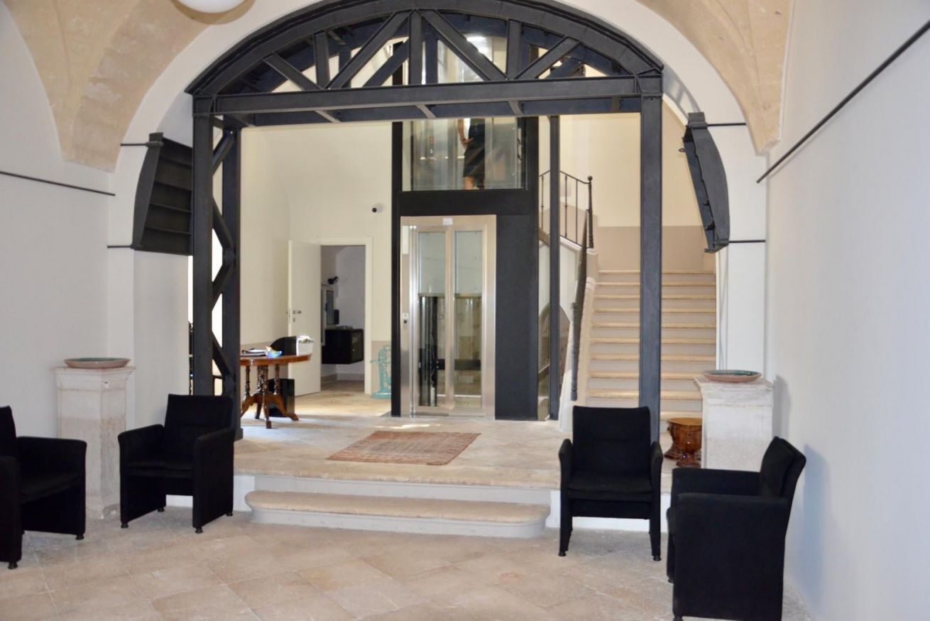 Un ampio interno con un'imponente porta ad arco che conduce a una scala. Si notano due poltrone nere e un tavolino con una ciotola su un piedistallo.
