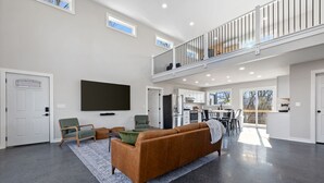 A spacious living room with a high ceiling, a large flat-screen TV, a brown sofa, and a dining area with a table and chairs. - Barndominium w/ Barrel Sauna, Mountain Views, King Bed & Fast WiFi (Berkeley Springs)