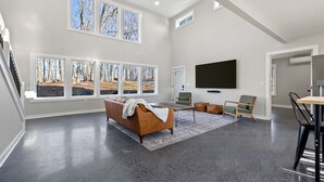 A spacious living room with a large window, a sofa, a coffee table, and a flat-screen TV. - Barndominium w/ Barrel Sauna, Mountain Views, King Bed & Fast WiFi (Berkeley Springs)