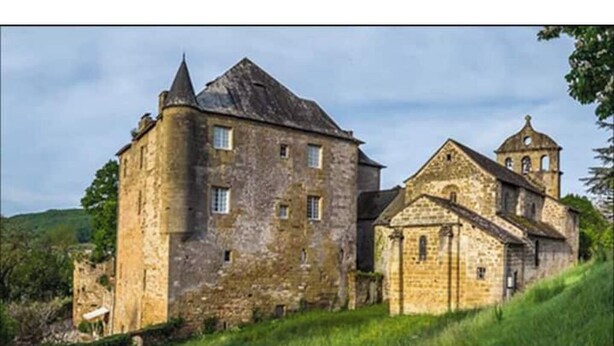 Un edificio histórico de piedra con una torre destacada y una estructura más pequeña adosada, presumiblemente una iglesia, en un paisaje verde y exuberante.