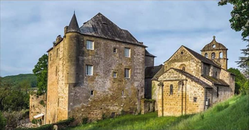 A historic stone building with a prominent tower and a smaller attached structure, likely a church, set in a lush, green landscape.