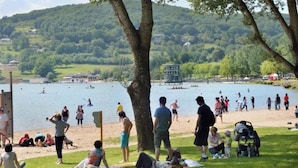 Un parque junto al lago con gente descansando sobre el césped, una torre de salvavidas y embarcaciones en el agua.