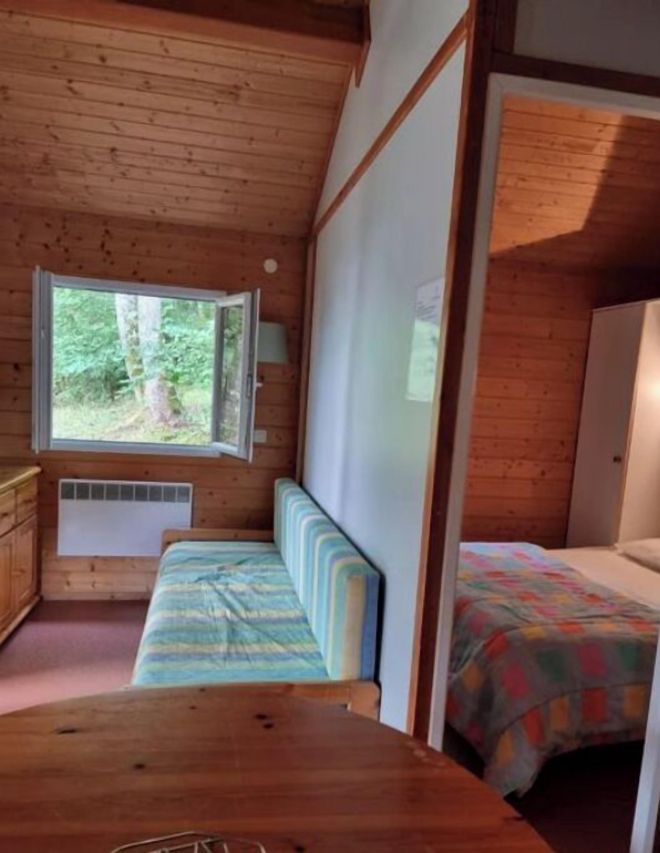 A cabin interior with a wooden ceiling, a window overlooking a forest, a bed with a colorful bedspread, and a small table.