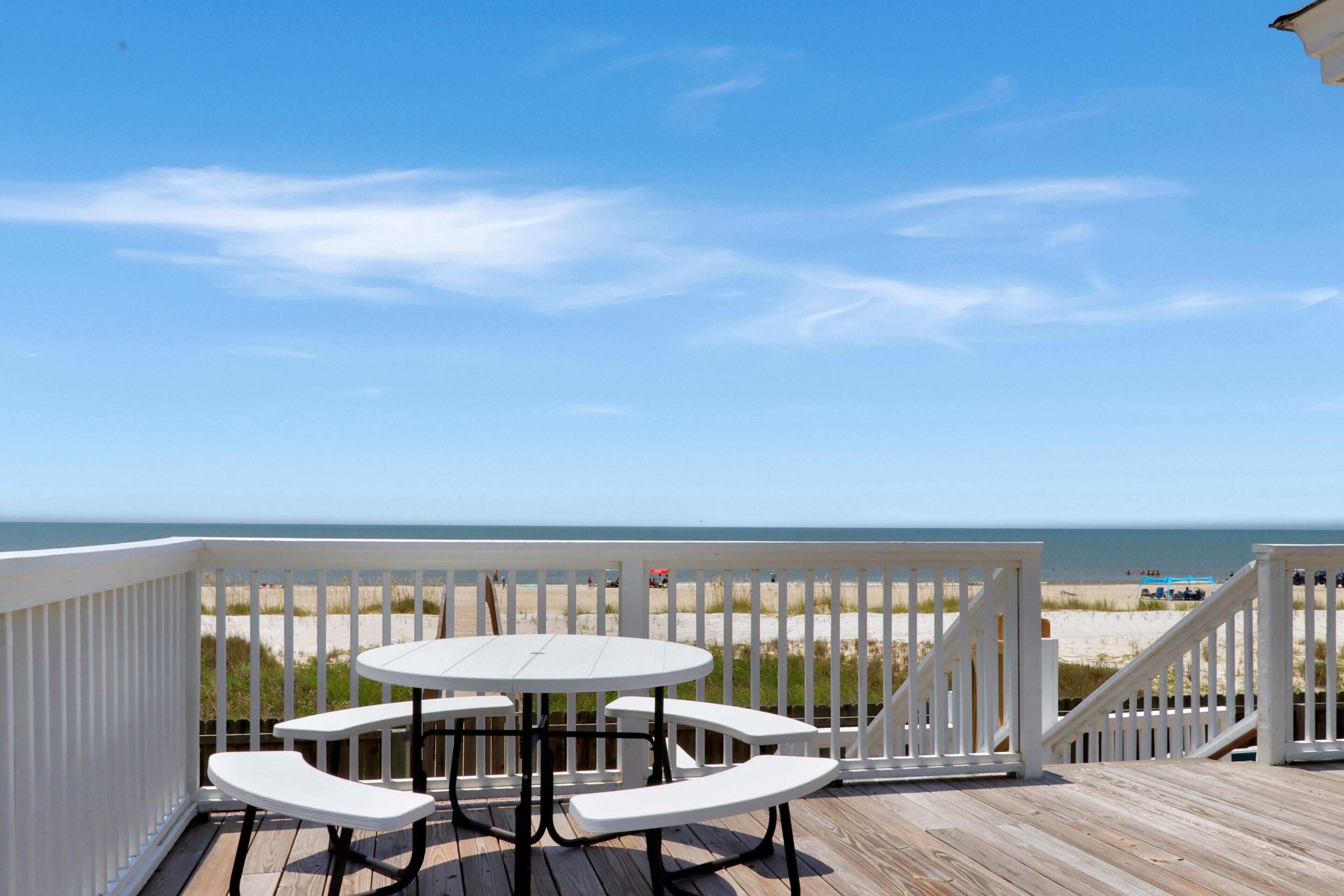 Une terrasse en bois avec une table et des bancs blancs, donnant sur une plage délimitée par une clôture et offrant une vue lointaine sur l’océan.