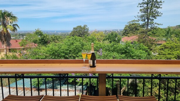 A balcony with a wooden table and chairs, overlooking a lush green landscape and a distant cityscape. - Enjoy Sydney's Northern Beaches. Unique district views, 1950s classic, sleeps 5 (Seaforth)