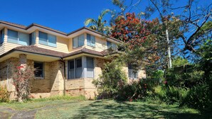 A two-story house with a brick facade, large windows, and a well-maintained garden with various plants and a tree with red flowers. - Enjoy Sydney's Northern Beaches. Unique district views, 1950s classic, sleeps 5 (Seaforth)