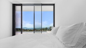 A bedroom with a large bed, white bedding, and a view of a landscape through a sliding glass door.