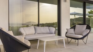 A modern outdoor seating area with a sofa and chairs, a white table, and large windows offering a view of the landscape.