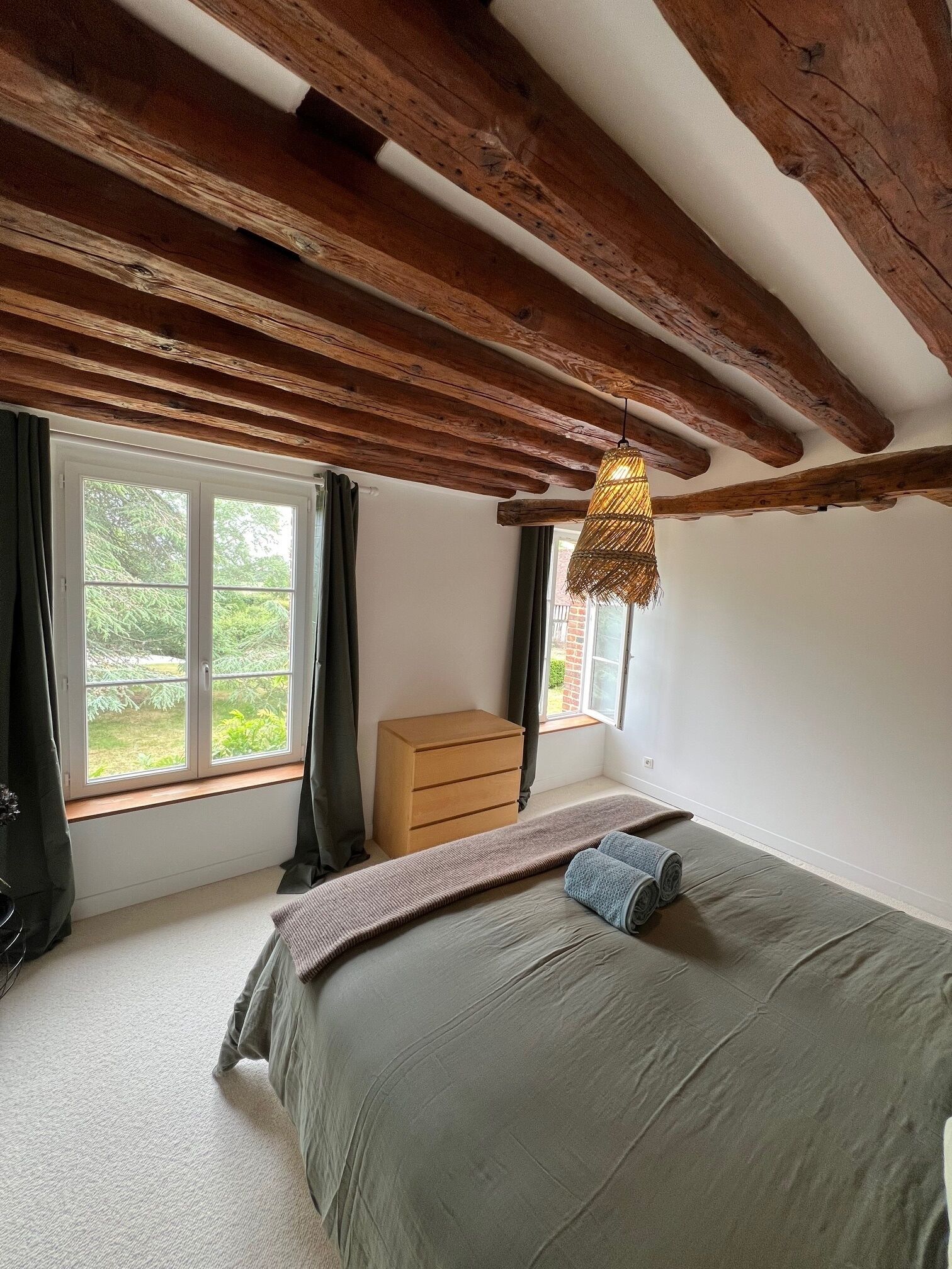 Un dormitorio con una cama, una ventana con cortinas, un aparador de madera y una lámpara colgante.