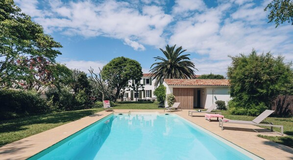 Una piscina con lettini, un piccolo edificio e una casa grande con tetto in tegole sullo sfondo.