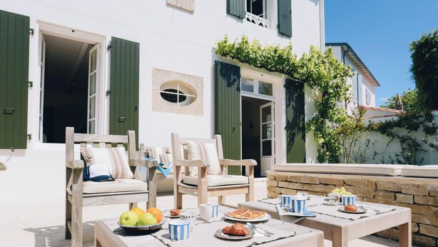 A white building with green shutters, outdoor seating with wooden chairs and cushions, a table set with breakfast items, and a stone wall. - Villa Phoenix - Ancient and modern charm (La Couarde sur mer)