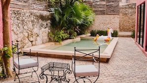 Pequena área de piscina ao ar livre, com duas cadeiras de metal e uma mesa, cercada por plantas e um muro de pedra.