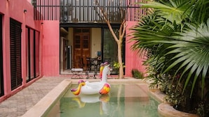 Piscina com bóia de unicórnio, rodeada por um edifício rosa e vegetação.