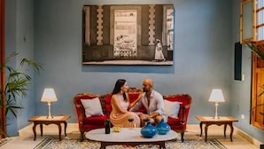 Um quarto com um sofá vermelho, duas mesinhas de cabeceira com abajures, um tapete estampado e uma pintura grande na parede.
