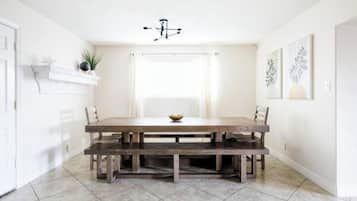 Un comedor con una mesa de madera y bancos, una pared blanca con estante y una ventana con cortinas.