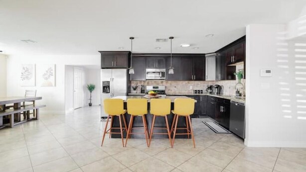 A modern kitchen with dark cabinets, a central island with yellow chairs, and a dining area with a table and chairs. - Rancho Relaxo: Dog friendly! Enjoy heated pool, hot tub, pool table, outdoor TV! (Scottsdale)