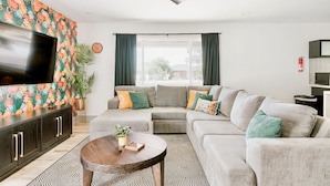 A modern living room with a sectional sofa, a flat-screen TV, a ceiling fan, and a large window with curtains.