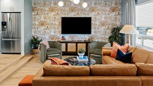 Een moderne woonkamer met een grote bank, een salontafel en een flatscreen televisie aan de muur. Er is een keuken met een koelkast en een eettafel met stoelen.