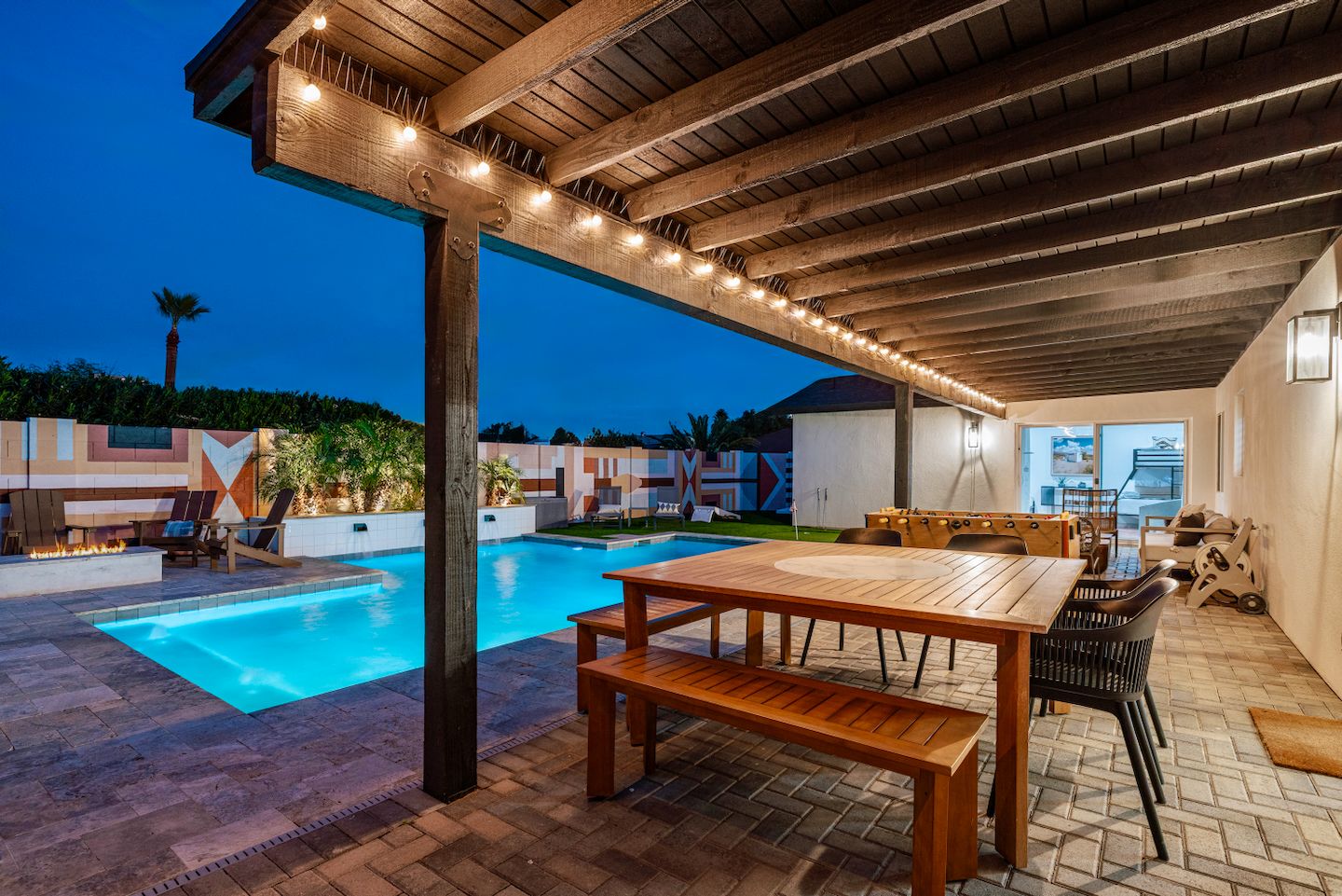 Área de piscina externa com pergolado de madeira, uma mesa e cadeiras, cercada por um deck revestido com azulejos.