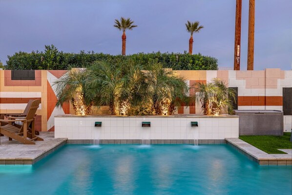 Una piscina con borde de baldosas, rodeada de palmeras y un muro decorativo con diseños geométricos.