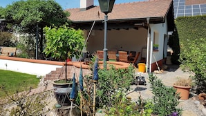 A house with a tiled roof, a chimney, a covered patio with chairs, and a garden with various plants and a bird feeder.