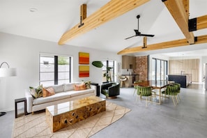 A spacious living area with a sofa, coffee table, and dining table. There is a kitchen area with a brick wall and wooden beams. - CoroMODo! Firepit, Outdoor kitchen (Phoenix)