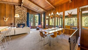 A spacious room with a stone fireplace, wooden ceiling, and large windows offering a forest view. - Trapper Cabin in Alpine Meadows w/ hot tub (Alpine Meadows)