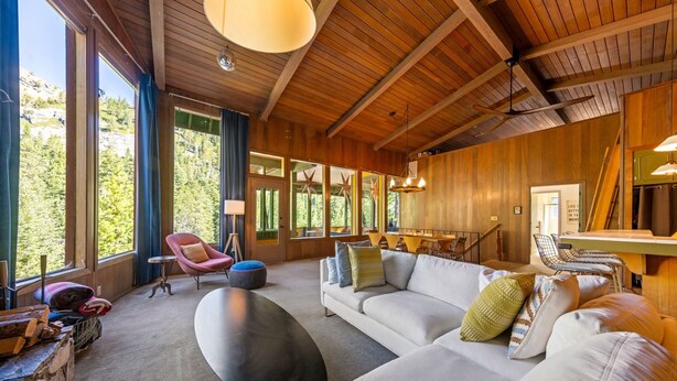 A spacious living area with a large sofa, a coffee table, and a dining table. There are large windows offering a view of the outdoors. - Trapper Cabin in Alpine Meadows w/ hot tub (Alpine Meadows)