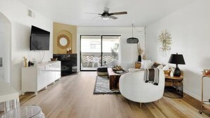 A modern living room with a white sofa, a coffee table, and a TV mounted on the wall. There is a fireplace, a ceiling fan, and a dining area visible through a glass door.