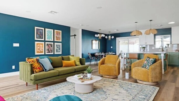 A modern living room with a green sofa, a coffee table, and a blue ottoman. There is a kitchen area with a dining table and chairs visible in the background.