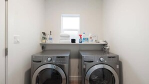 A laundry room with two front-loading washing machines, a shelf with cleaning supplies, and a window above the machines.