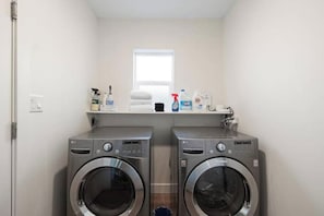 A laundry room with two front-loading washing machines, a shelf with cleaning supplies, and a window above the machines.