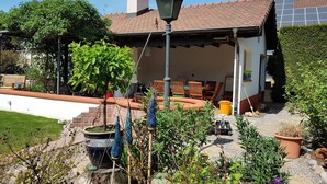 Una casa con techo de tejas, chimenea, un porche cubierto con sillas y un jardín con diversas plantas y un comedero para pájaros.