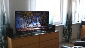 Una sala de estar con un televisor de pantalla plana sobre un mueble de madera, un sofá, una mesita con un control remoto y una ventana con cortinas.