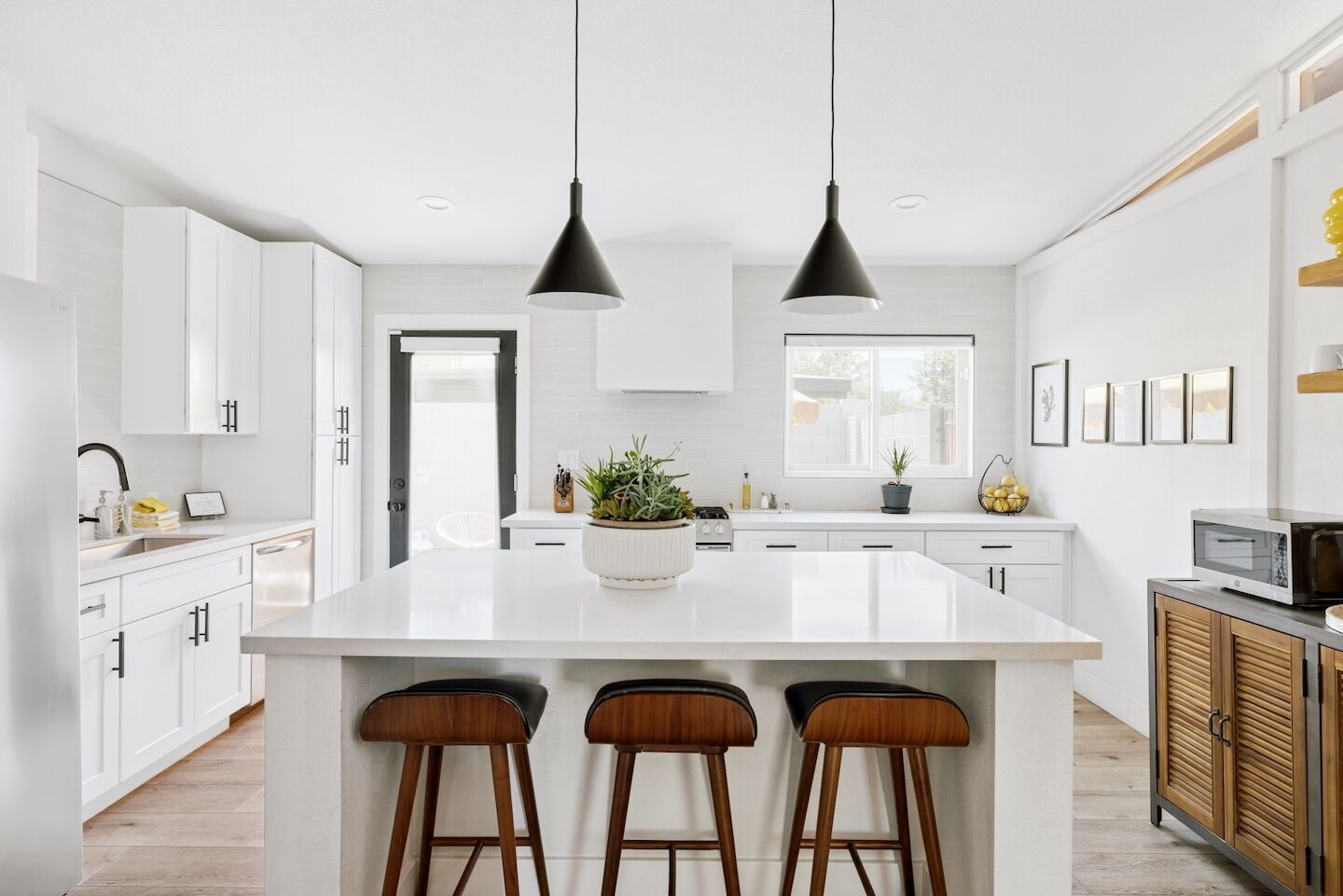 Une cuisine moderne avec des meubles blancs, un îlot central doté de tabourets, un éclairage suspendu et une vue sur l’extérieur.