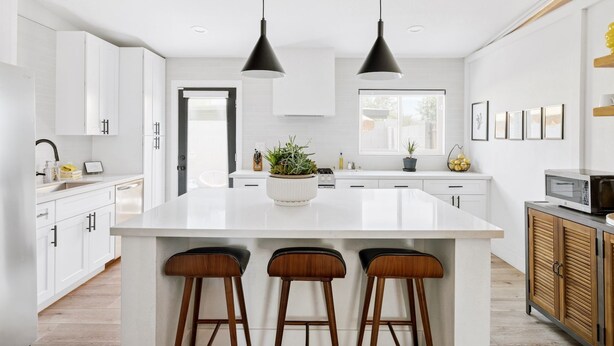 Una cocina moderna con armarios blancos, una isla central con taburetes, iluminación colgante y vistas al exterior.