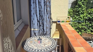 A small outdoor patio with a tiled table and a single chair, a window with a curtain, and a wall with climbing plants. - Sweet Escape. T1 BIS ROMANTIC (Bourg-lès-Valence)