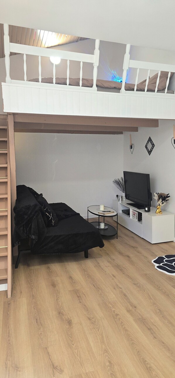 A loft bedroom with a bunk bed, a black sofa, a small round table, a TV on a white cabinet, and a wooden floor.
