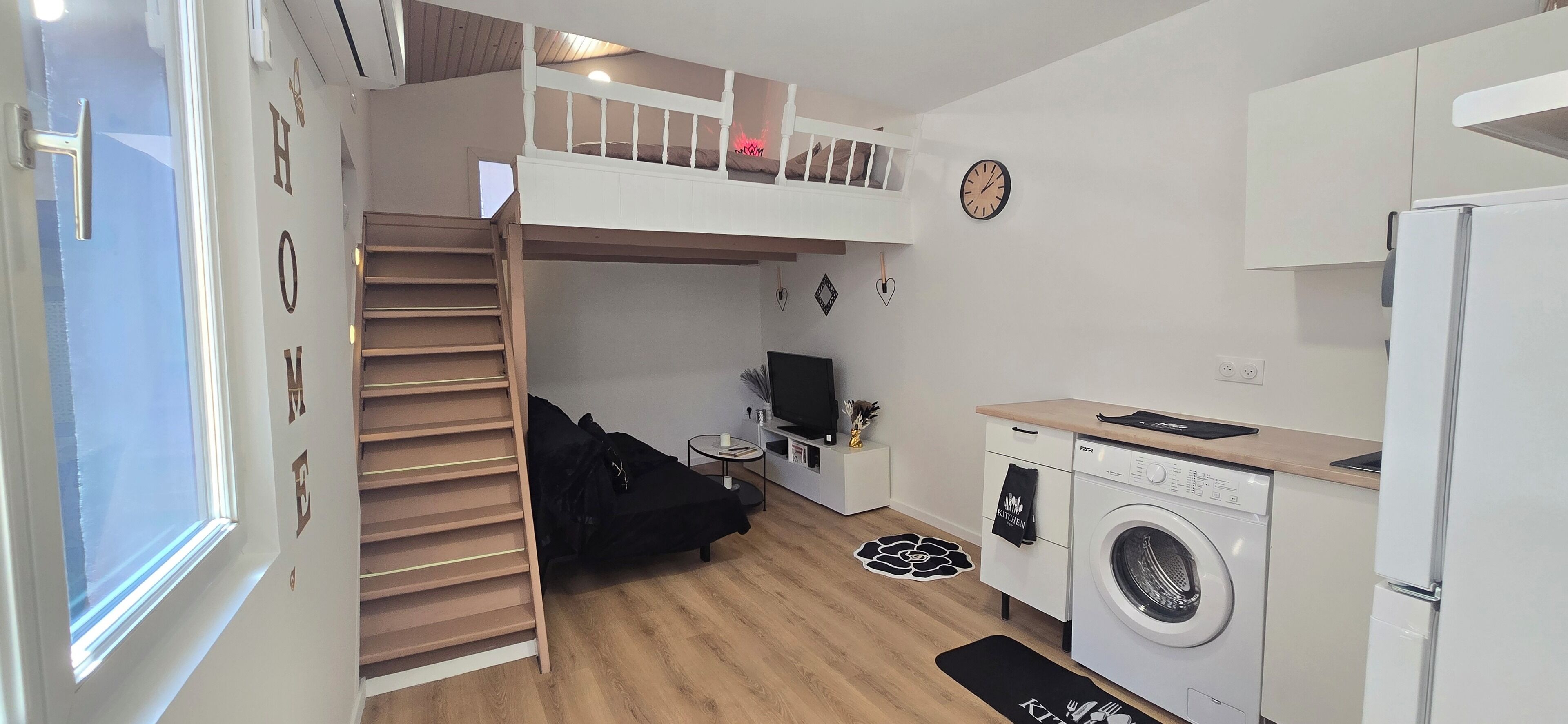 Une cuisine compacte équipée d’un lave-linge, un coin repas avec une table et des chaises, un lit superposé avec un téléviseur et une horloge au mur.