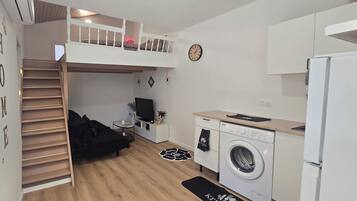 Une cuisine compacte équipée d’un lave-linge, un coin repas avec une table et des chaises, un lit superposé avec un téléviseur et une horloge au mur.