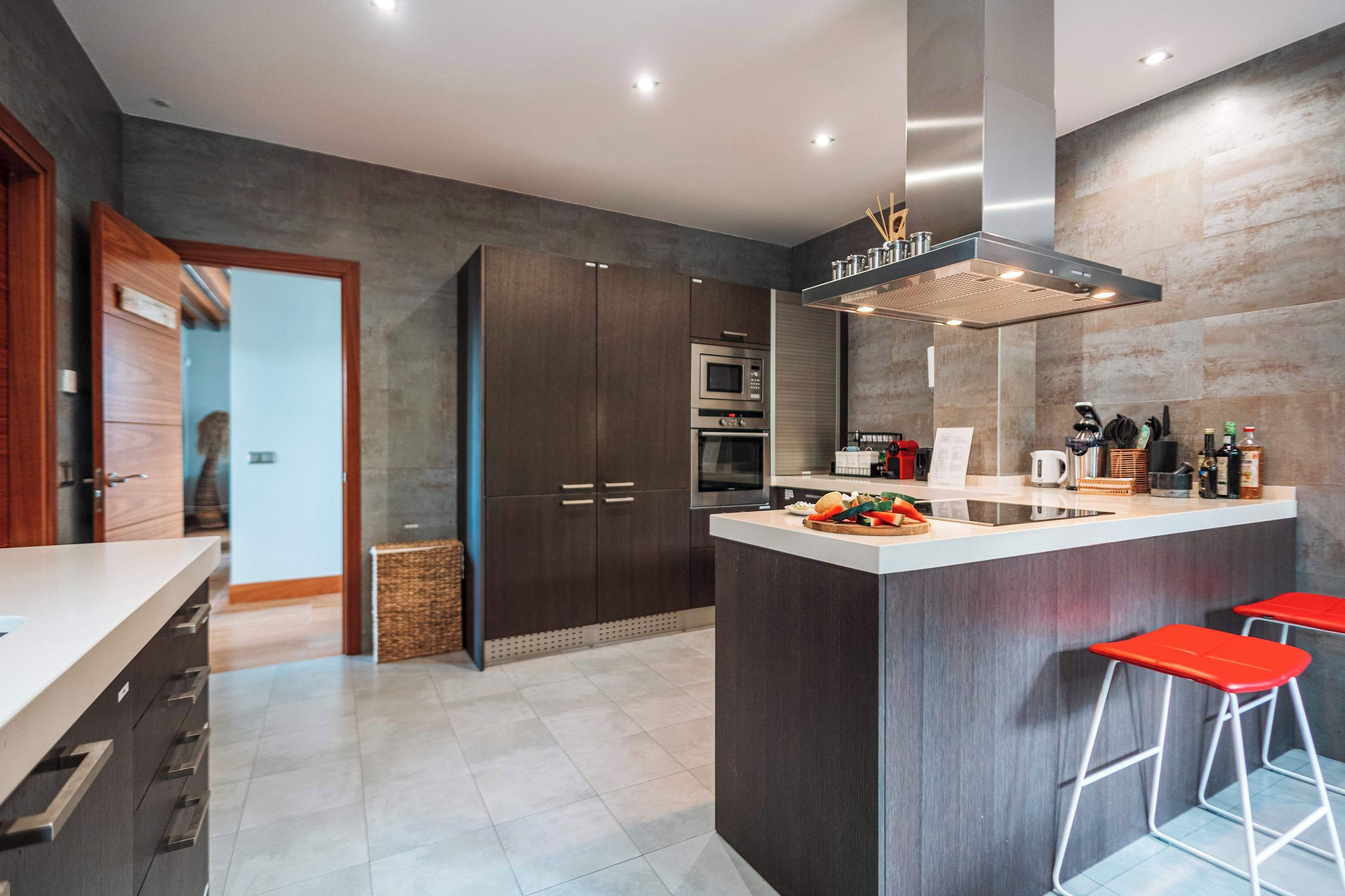 A modern kitchen with dark cabinetry, a central island with stools, stainless steel appliances, and a large range hood.