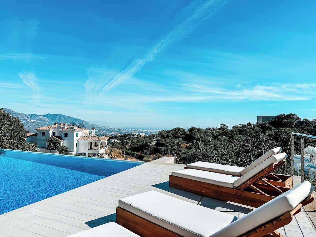 A swimming pool with lounge chairs, a wooden deck, and a view of a white building and mountains.