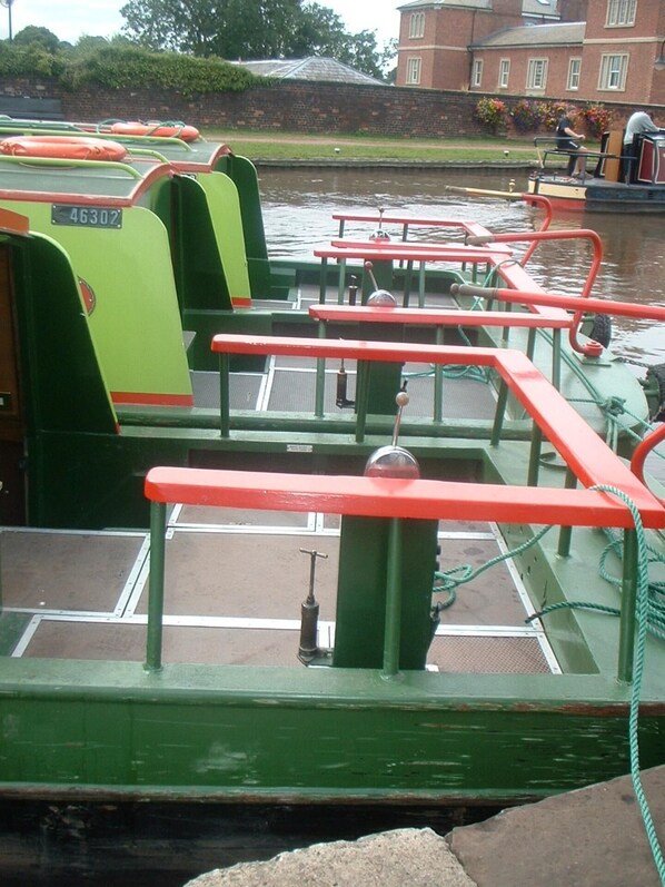 Un bote verde con barandales y bancos rojos, amarrado a un canal con edificios al fondo.