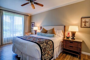 Una habitación de hotel con una cama grande, dos mesitas de noche con lámparas, un ventilador de techo y una ventana con cortinas traslúcidas.