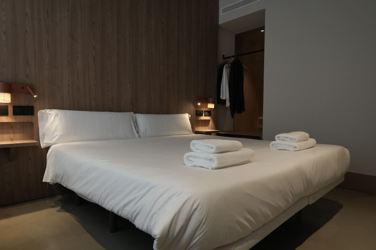 A hotel room with a large bed, a wooden headboard, a nightstand with a lamp, and a mirror with a towel rack.