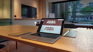 A laptop on a wooden table with a view of a cityscape through large windows. - CASA ROMA Hostel Boutique (Tarragona)