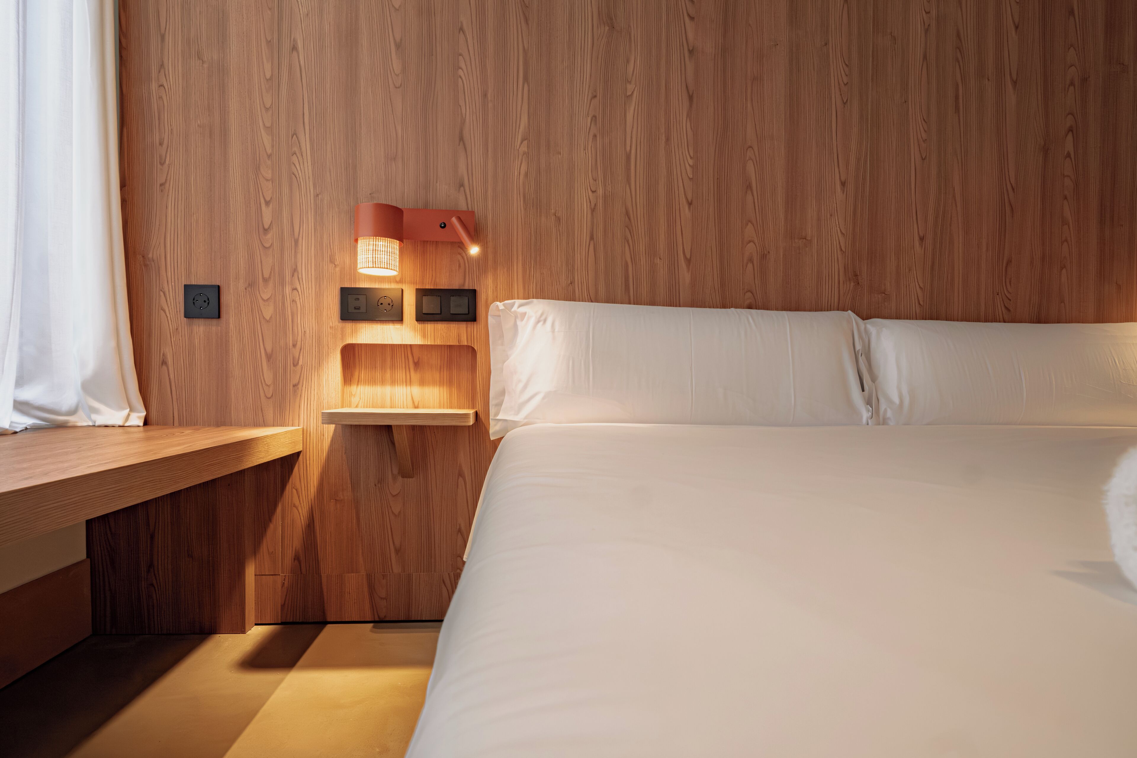A bedroom with a wooden headboard, a bedside table, a bed with white bedding, and a wall-mounted light fixture.
