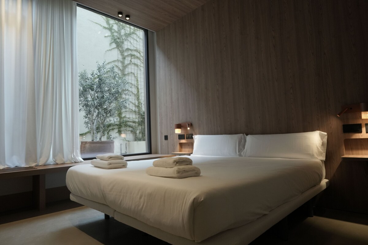 A modern hotel room with a large bed, a window with curtains, a bedside table with a lamp, and a view of trees outside.